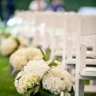 White floral arrangements along a row of white ceremony chairs outdoors
