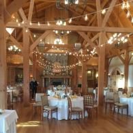 Elegant wooden banquet hall with round tables, white tablecloths, and string lights under exposed beams