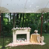 Outdoor party dessert table under a white canopy, with pink floral decor and a gold-draped side table.