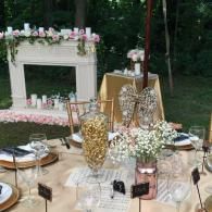 Elegant outdoor table setting with pink-and-white floral decor, gold accents, and clear chairs in a garden setting