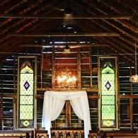 Dimly lit rustic church interior with stained-glass windows and a white-draped altar under a chandelier