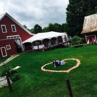 Barnyard scene with a person in a heart-shaped flower bed on green grass beside red barns and a white tent