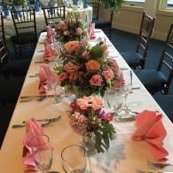Long banquet table with pink floral centerpieces, folded napkins, and glassware in a dining room
