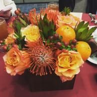 Orange and yellow flower arrangement with fruit in a black box on a table