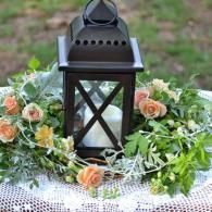 Black lantern centerpiece surrounded by peach roses and greenery on a lace tablecloth
