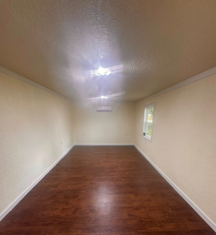 Empty room with wood floors, cream walls, white trim, ceiling lights, and a small window.