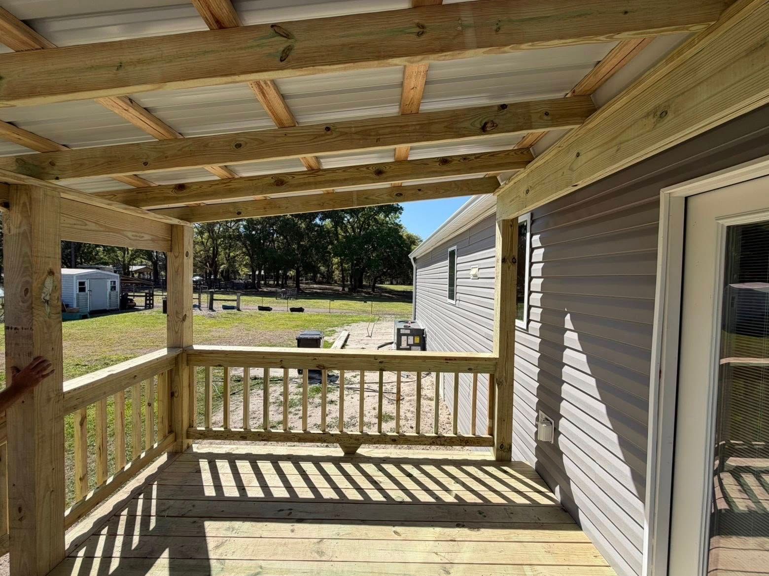 Covered wooden porch with railing beside a gray house, overlooking a grassy yard.