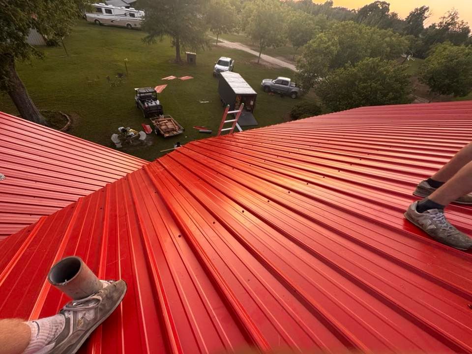 View from a red metal roof over a grassy yard with an RV, grill, and cars below.