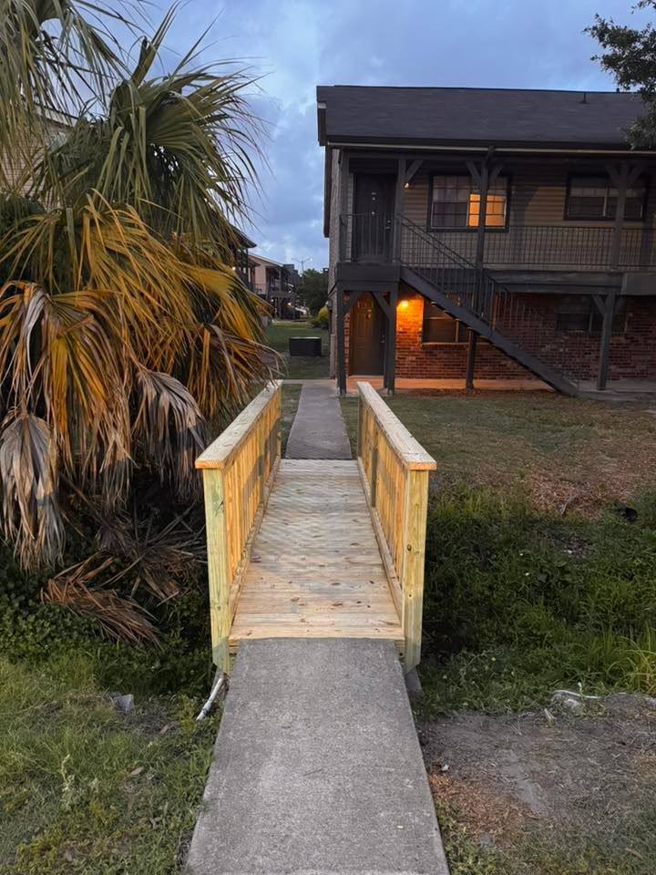Wooden ramp leading to a raised house at dusk, with palm trees and porch lights on.
