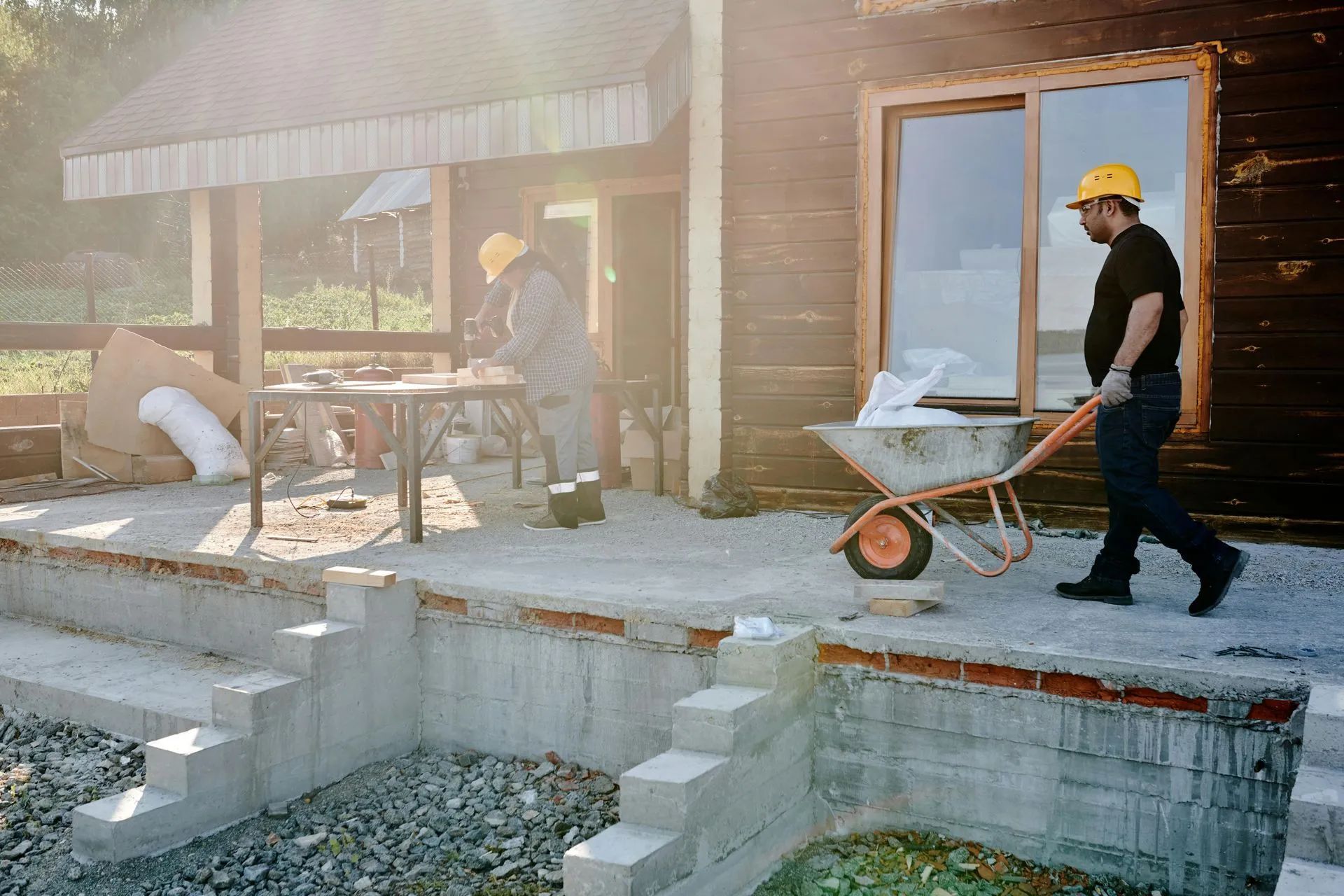 Two construction workers in yellow hard hats outside a wooden building. One prepares materials, the other pushes a wheelbarrow.