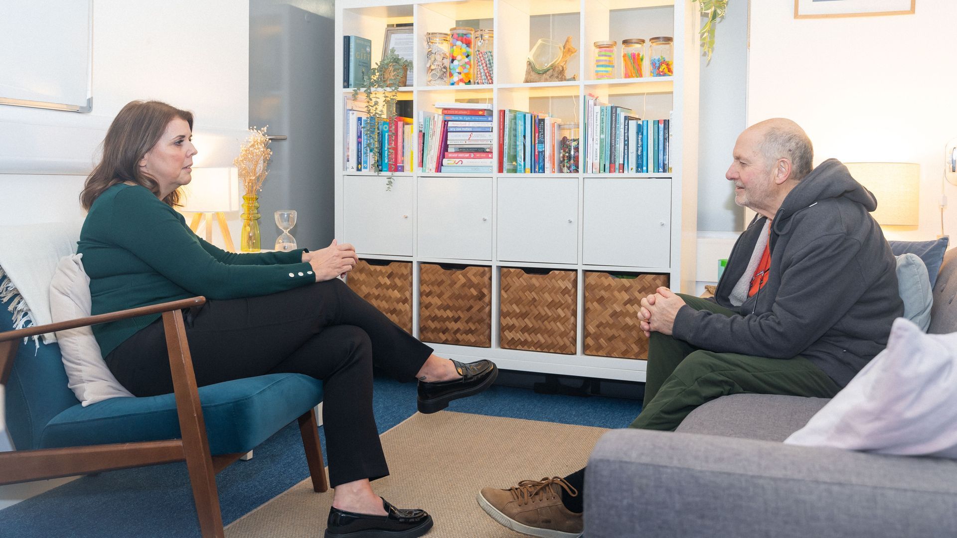 Woman in teal top and man in jacket converse in a room. Bookshelf and couch in the background.