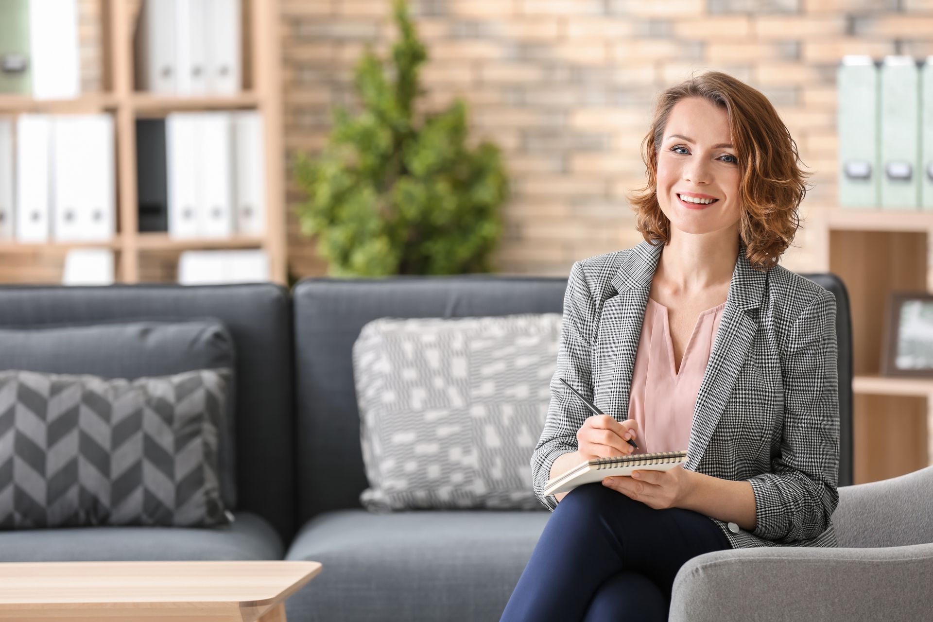 Woman in blazer sits in a chair, smiling while holding a notebook, in an office setting.
