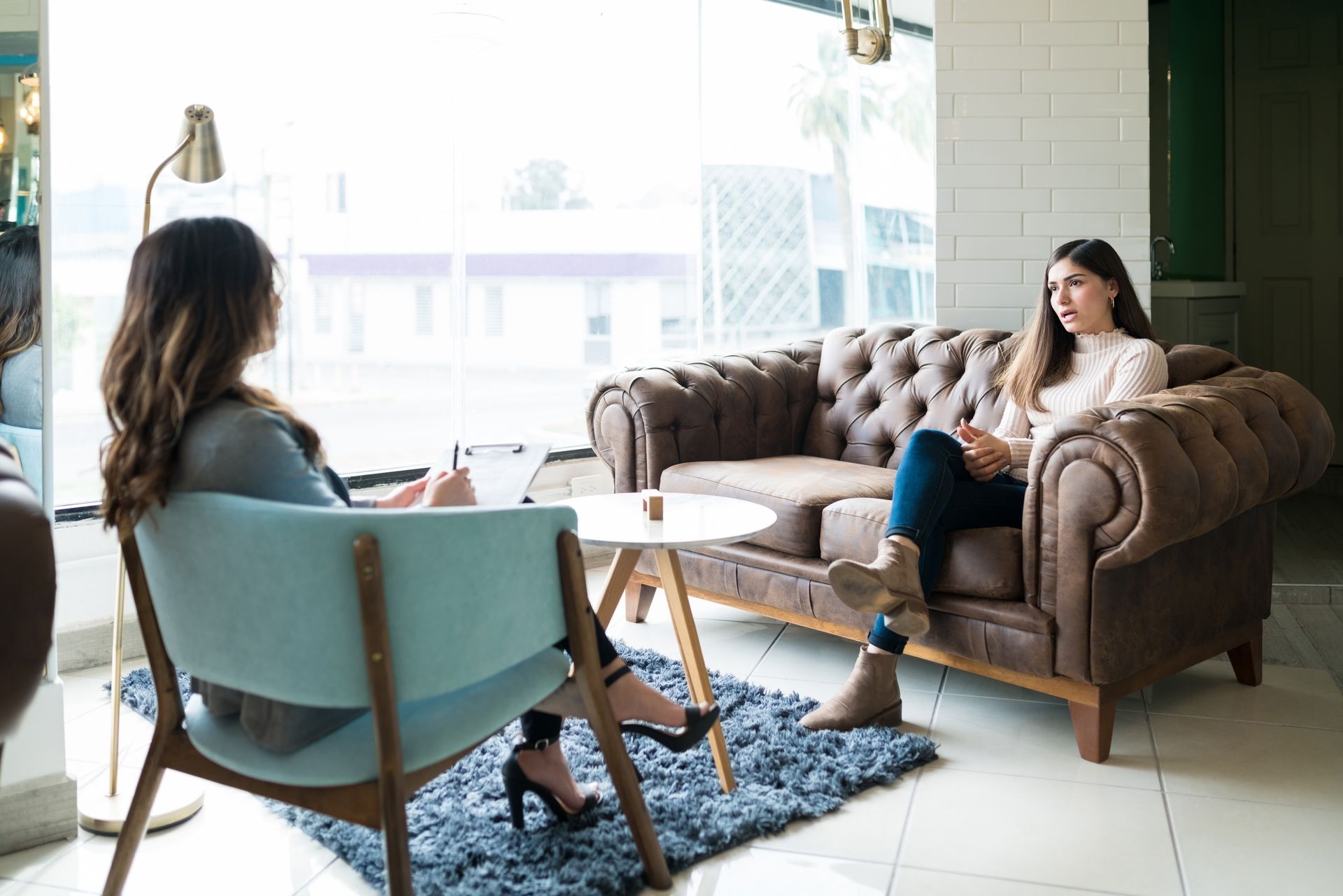 Woman seated in a chair, facing another woman on a sofa. They are in a counseling session indoors.