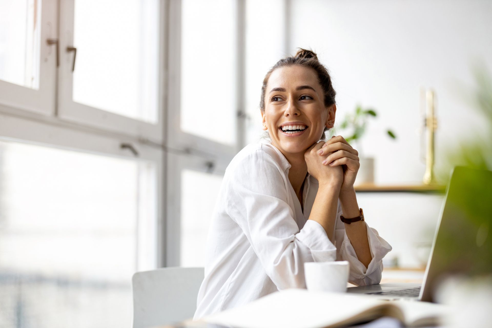 Woman smiles, leaning on hands, at desk near a window, with a laptop and coffee.