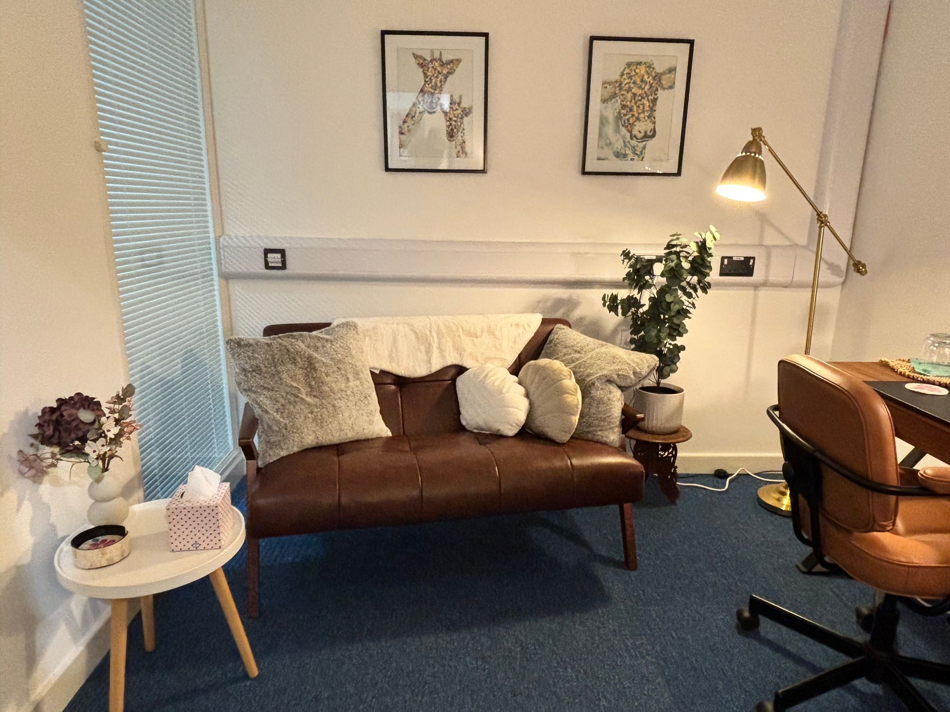 A cozy room with a brown couch, artwork, and a desk. Includes a gold lamp, a small table, and a plant.
