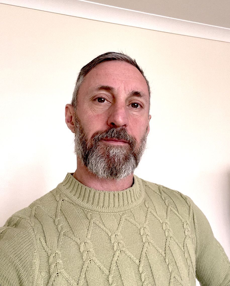 Woman smiles, wearing a patterned shirt and lanyard, in a room with a painting and greenery.