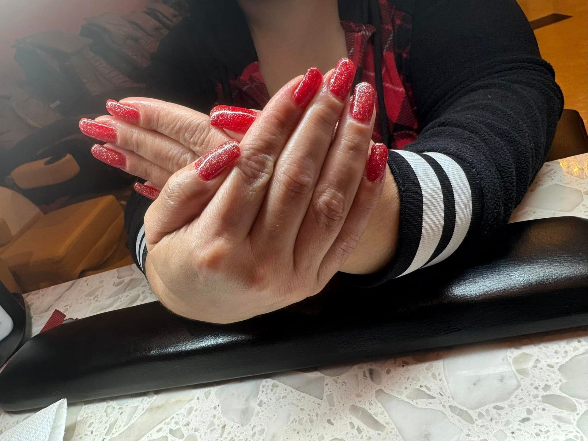 Hands with long, red, glittery acrylic nails resting on a black armrest.