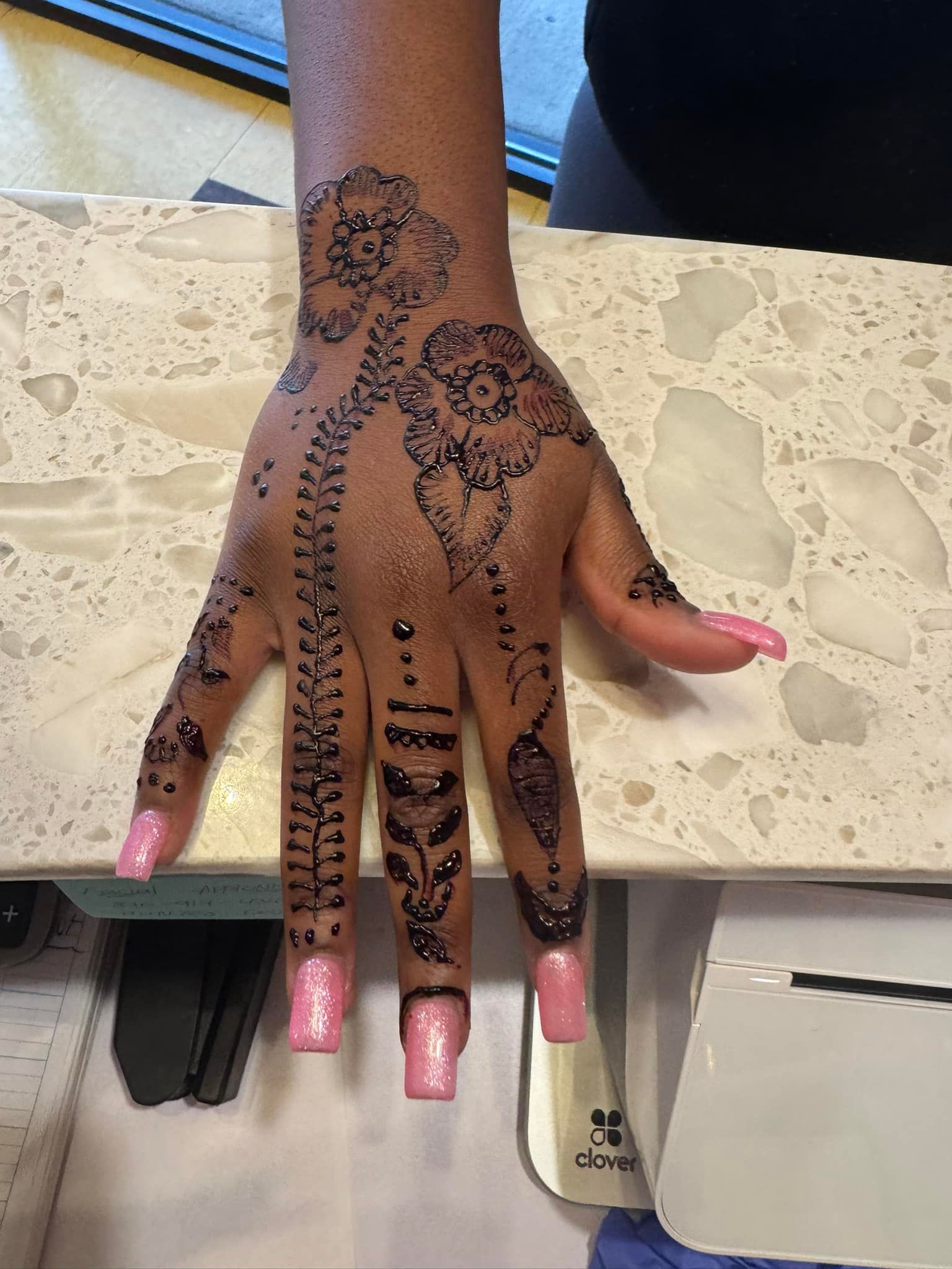 Hand with black henna design, long pink nails, resting on a beige counter.