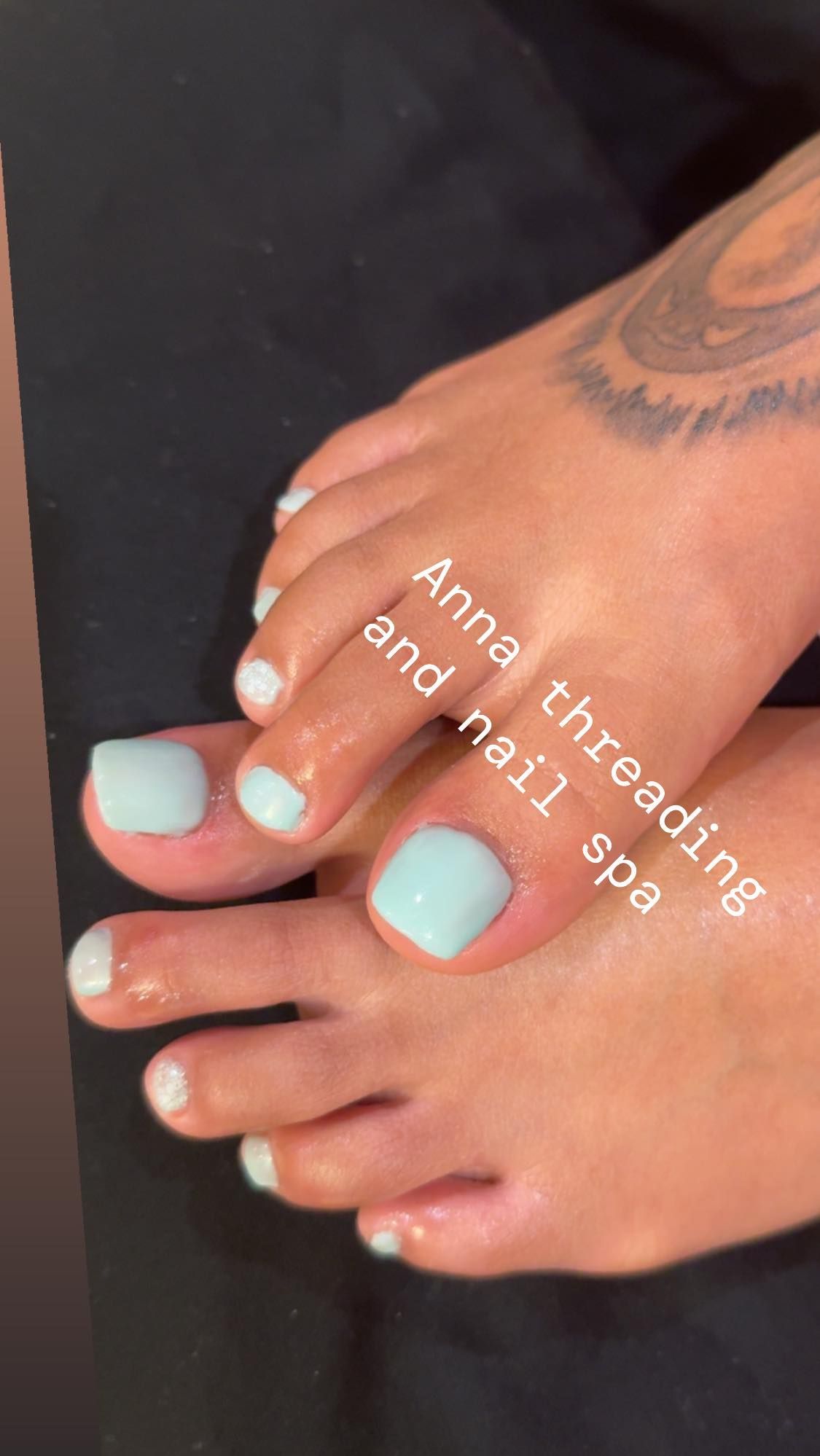 Feet with light blue and white toenail polish. Tattoo on the ankle.
