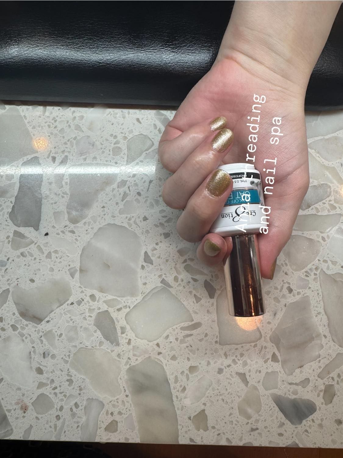Hand with gold nail polish holding a beauty product on a marble counter.