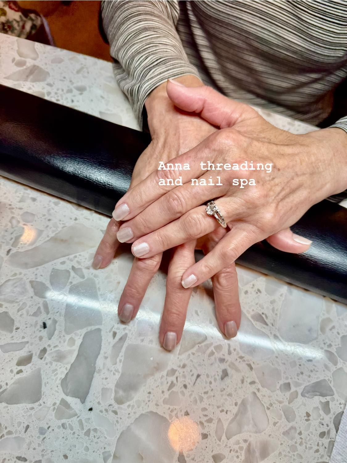 Hands with manicured nails on a marble surface. The hands are resting on a black armrest.