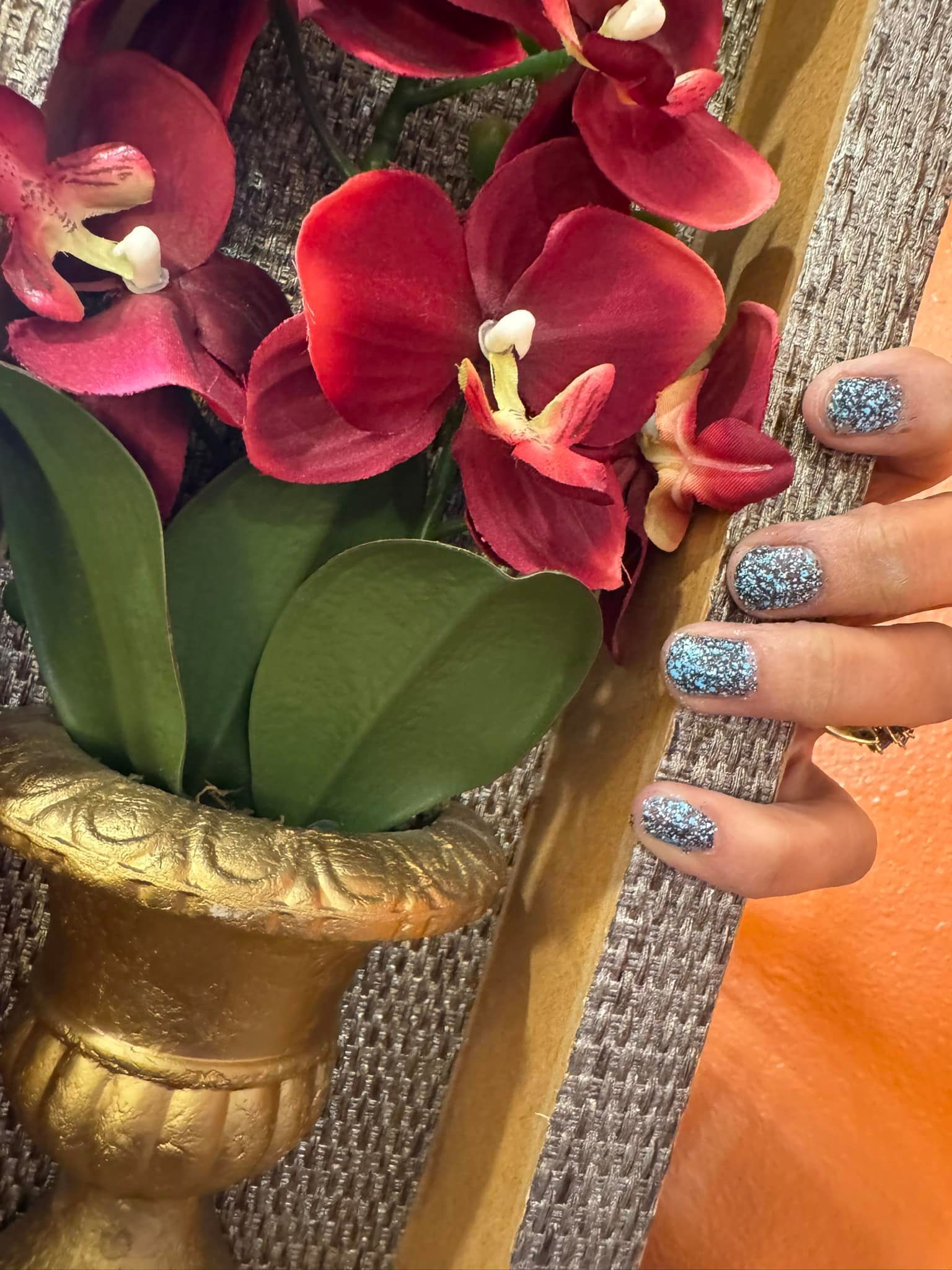 Hand with blue glitter nails next to a burgundy orchid in a gold pot.