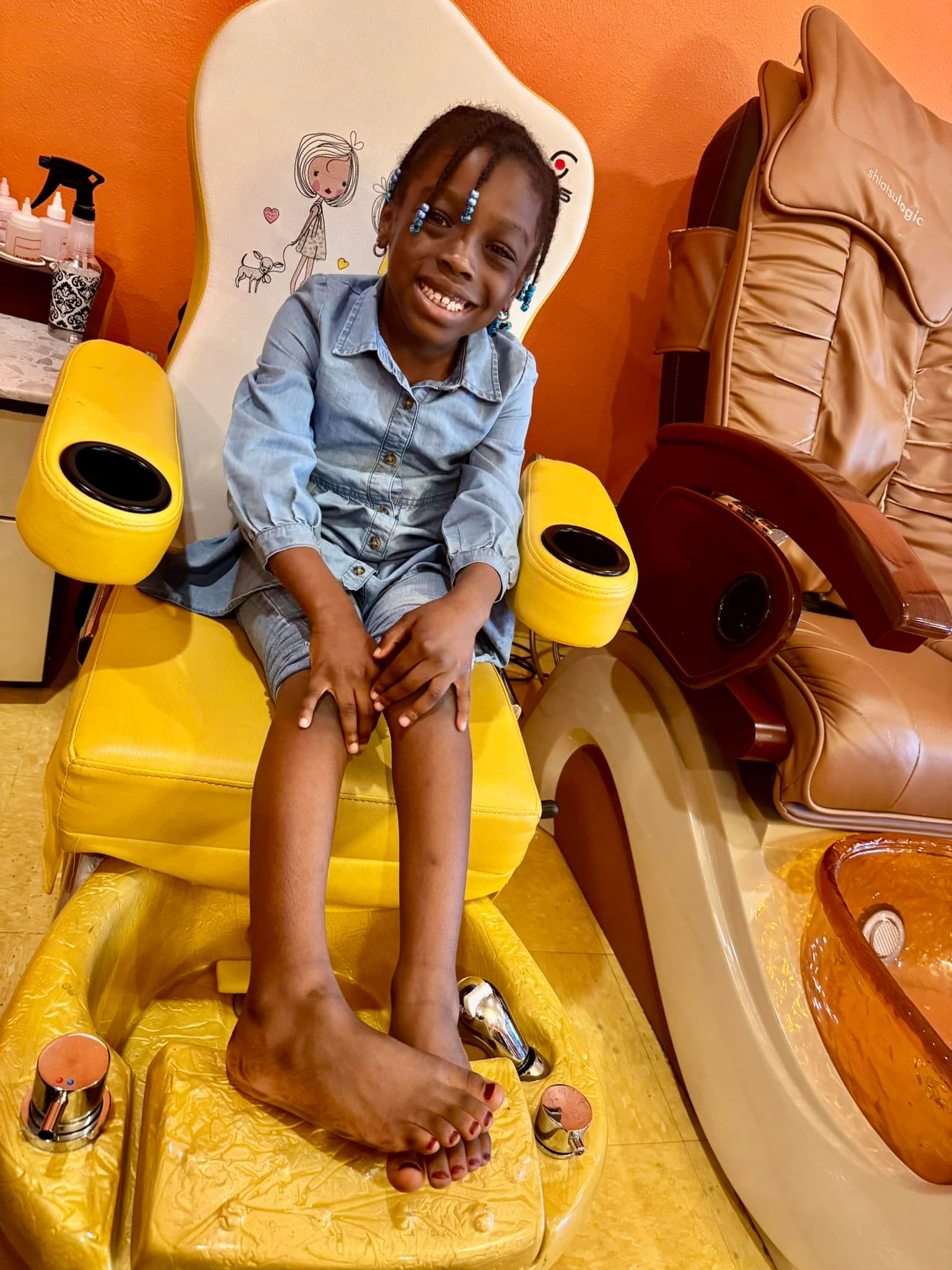 Girl smiling in a yellow pedicure chair, wearing denim, at a nail salon.