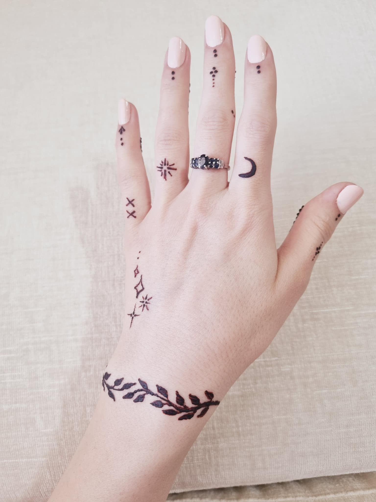 Hand with light pink nails, adorned with black henna tattoos of a crescent moon, stars, and leaf pattern.
