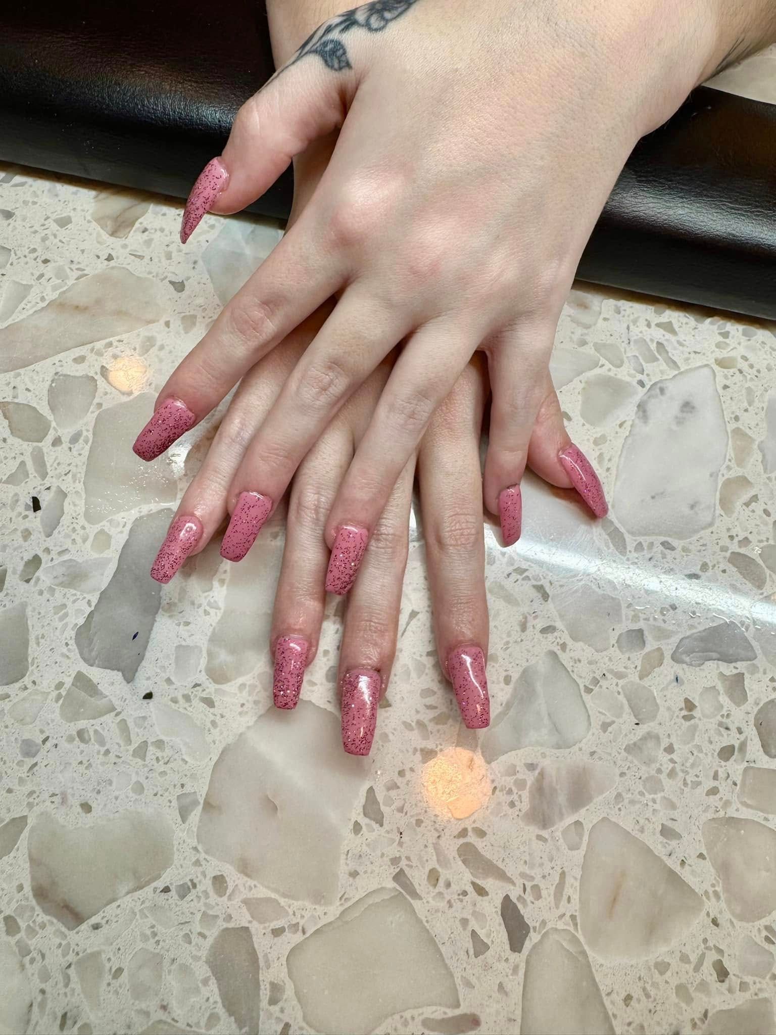 Hands with long, pink glitter nails on a speckled countertop, one hand has a wrist tattoo.