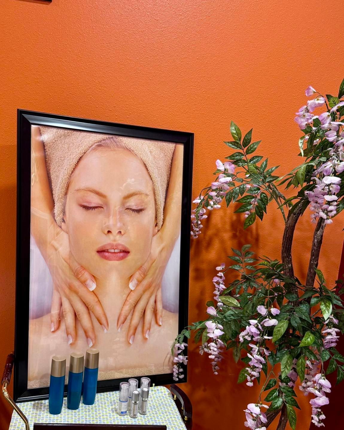 Spa display: framed photo of person receiving a facial treatment, skincare products, and flowering branch against an orange wall.