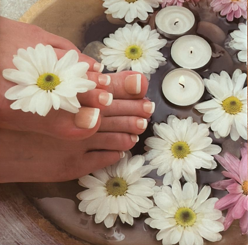 Foot with French-tip toenails in water with daisies, pebbles, and lit candles.