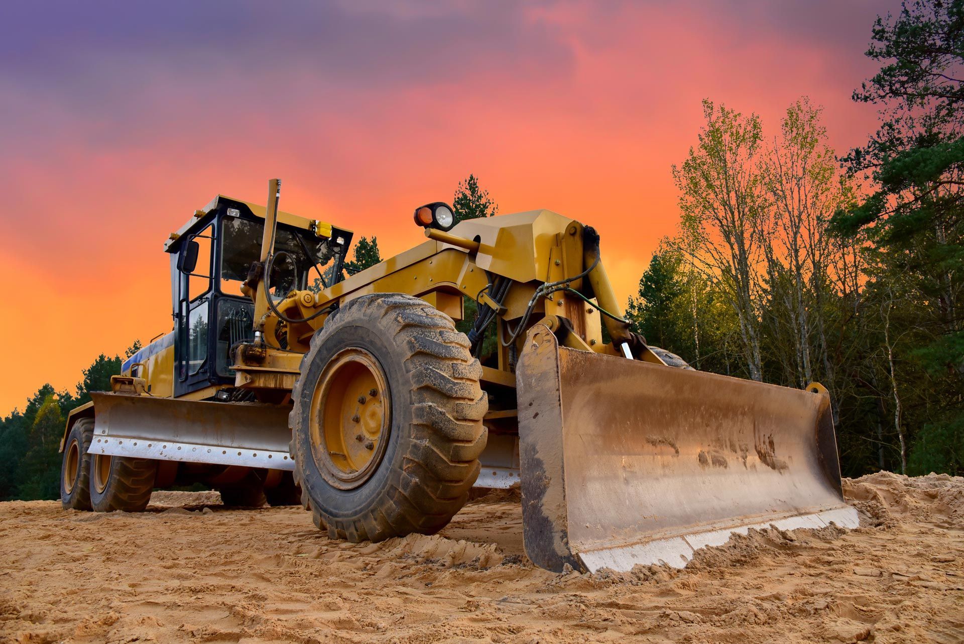 Yellow grader on dirt road against an orange and pink sunset; trees in the background.