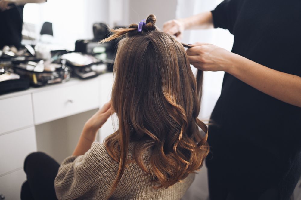 Hair stylist curling a client's brown hair with curling iron; vanity table and mirror in background.