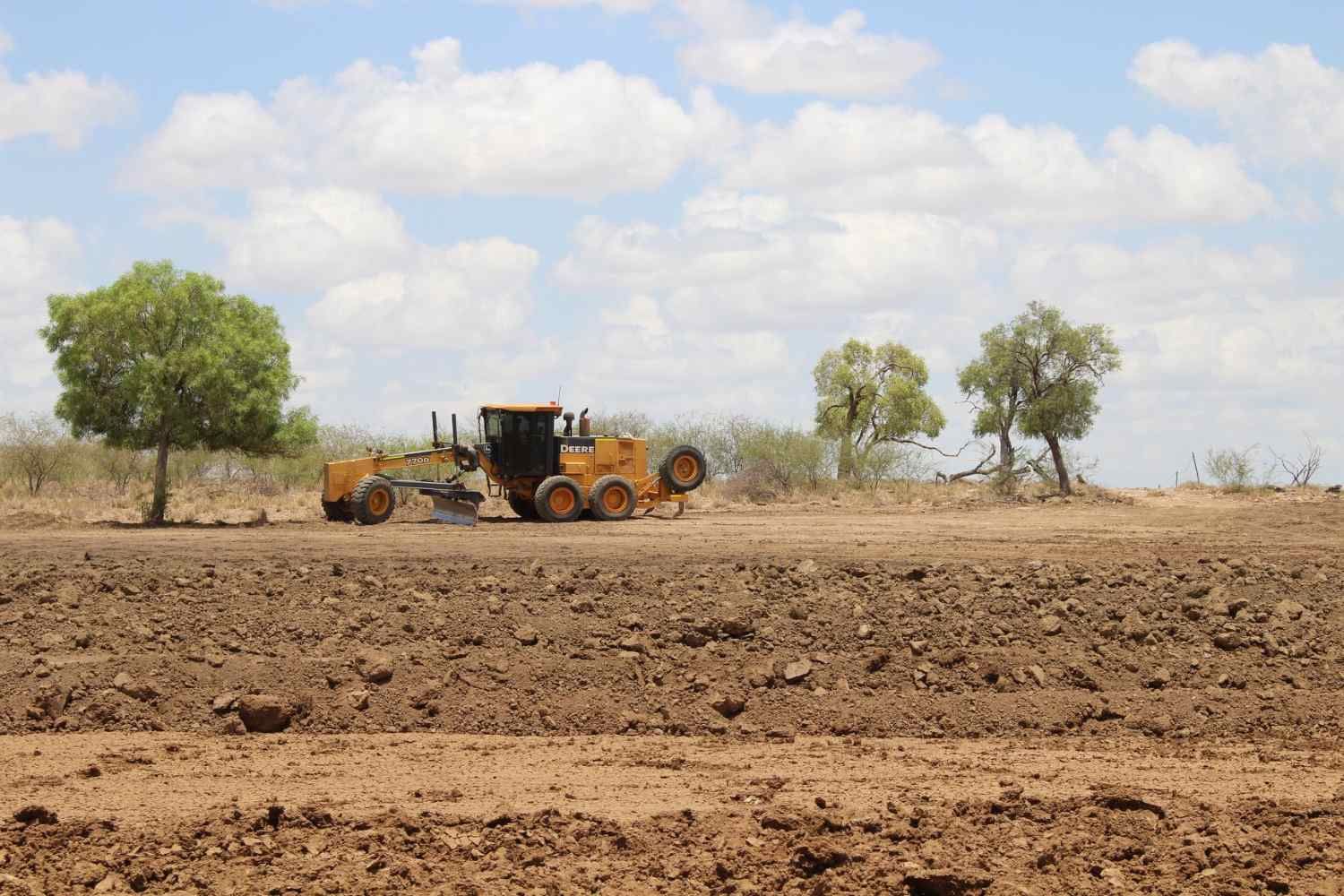Yellow Tractor is Plowing a Dirt Field With Trees in the Background — Burnett Contracting in Cloncurry, QLD