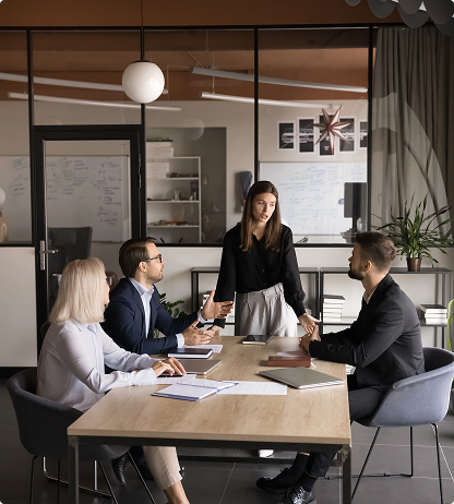 Office meeting: woman standing, gesturing, leading discussion with three colleagues seated at table.