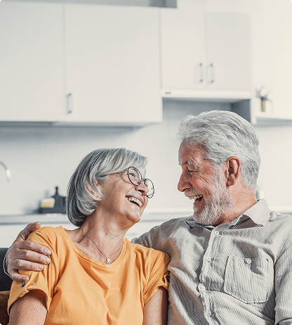 Smiling couple embracing, laughing indoors. Woman in yellow shirt, glasses; man in light shirt, gray hair.