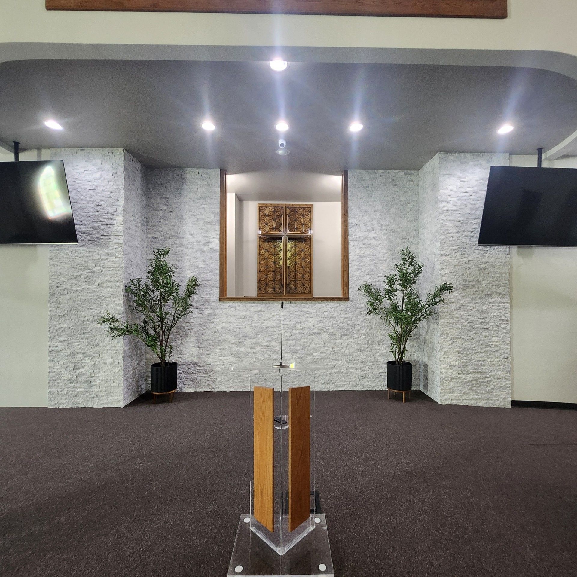 A church interior with stone walls, a wooden cross, plants, and a podium.