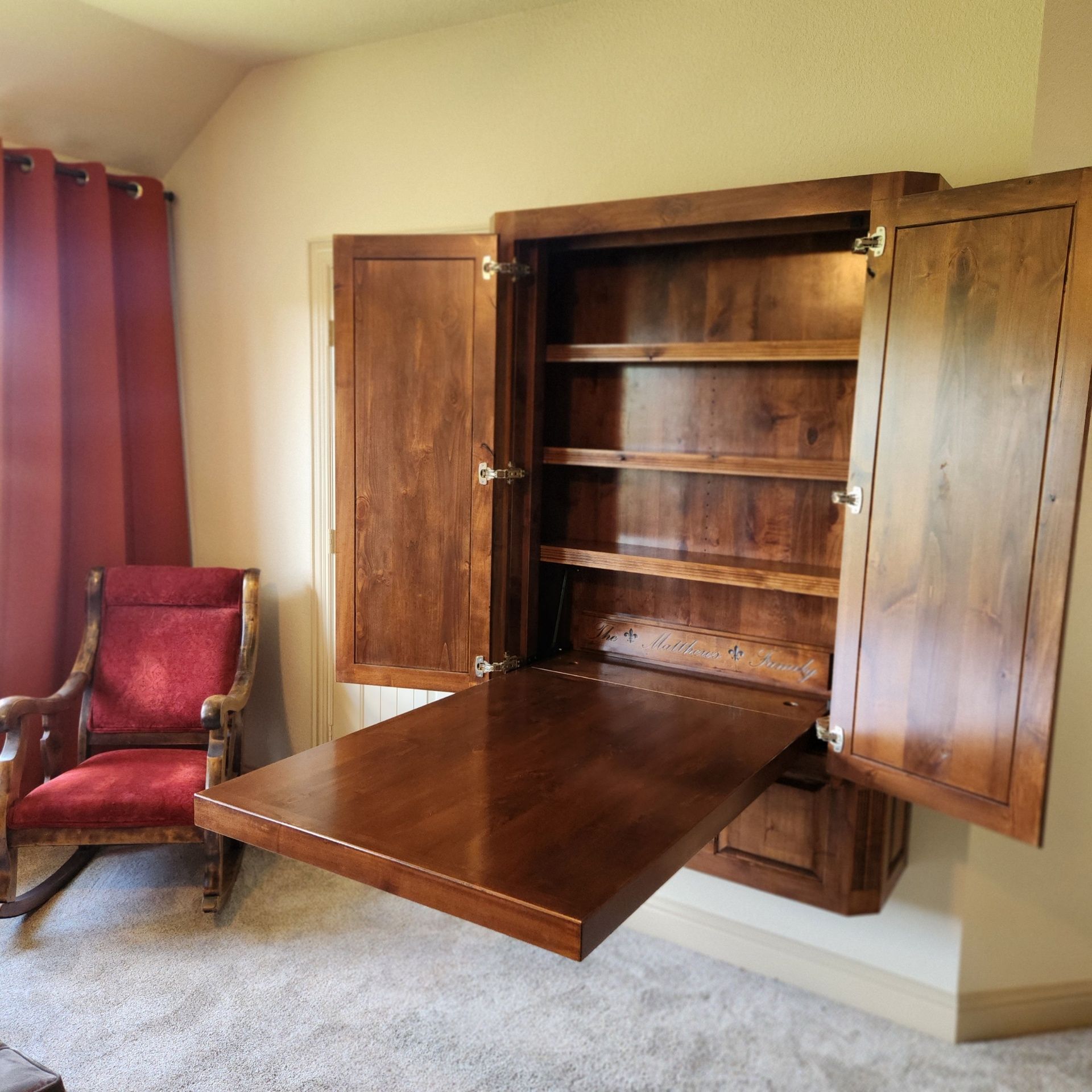 Wooden wall-mounted cabinet with open doors and a fold-down desk. Shelves inside. Chair and curtain to the left.