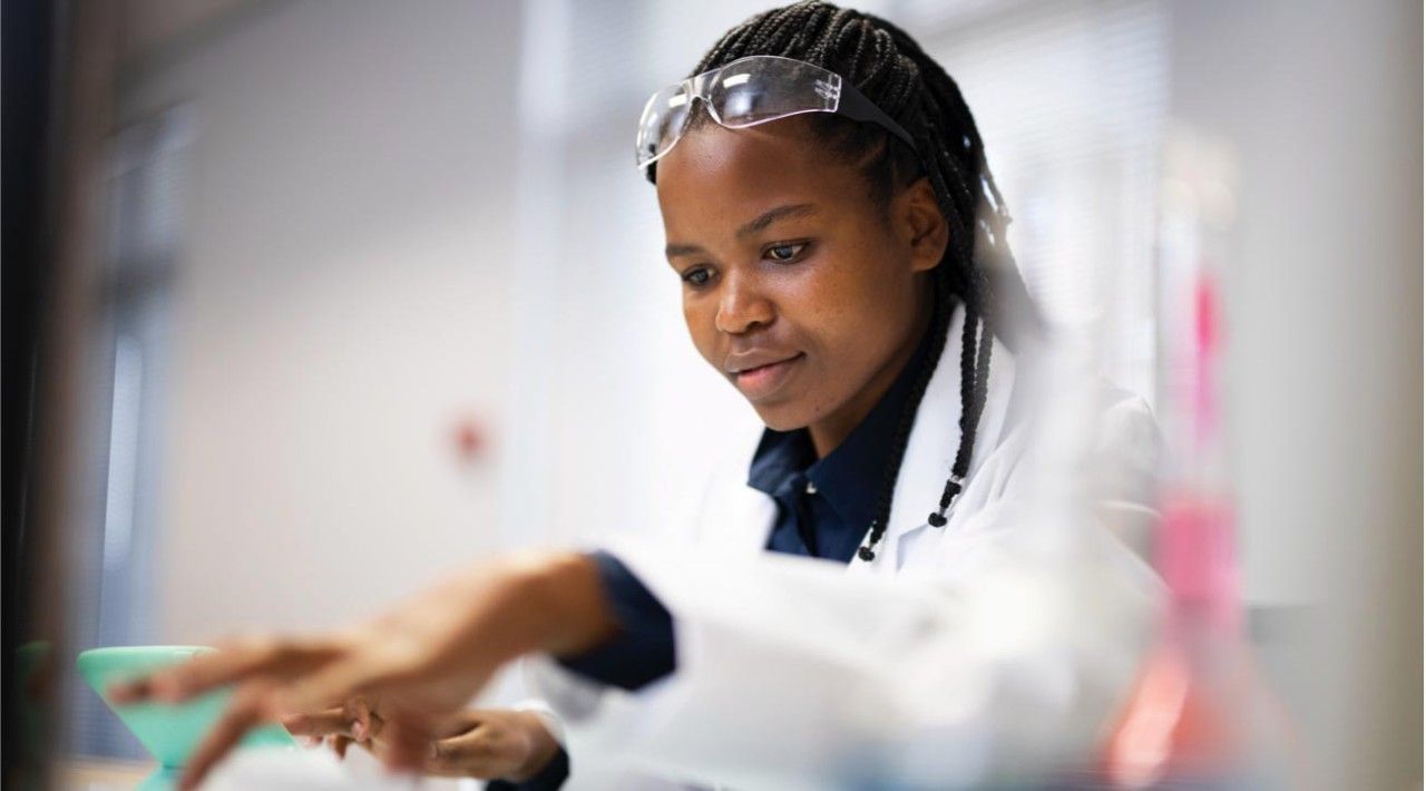 A woman in a lab coat and goggles is working in a lab.