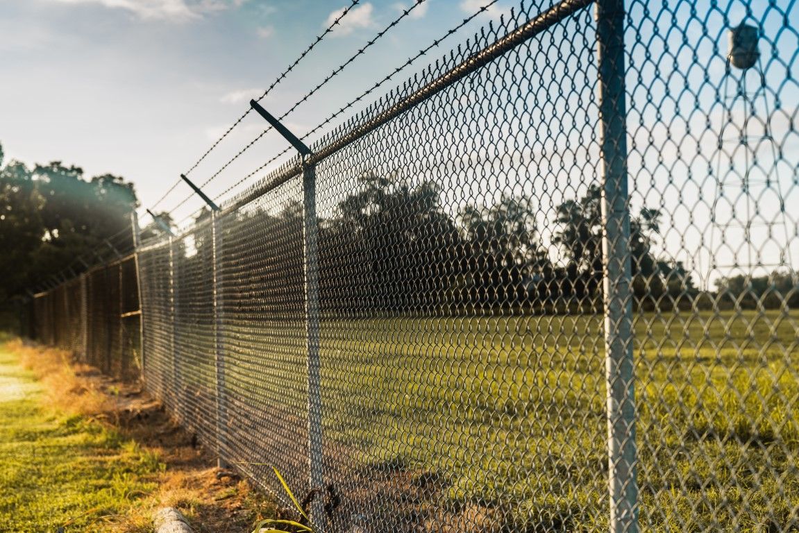 Chain-link fence with barbed wire on top, casting a shadow across a grassy field under a clear blue sky.