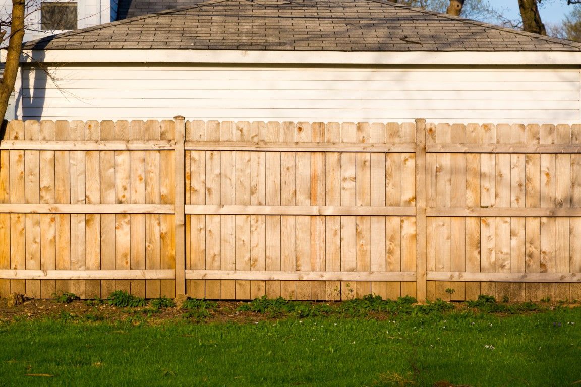Wooden picket fence with vertical slats, set against a backdrop of green trees under a clear blue sky. 
