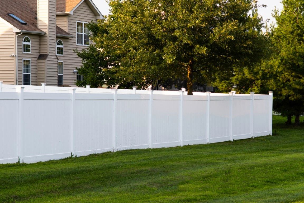 A white vinyl fence stands in front of green grass and a sidewalk, framed by tall trees.