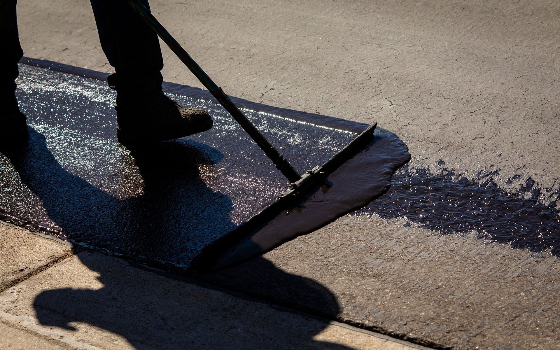 A person is spreading asphalt on a sidewalk with a broom.