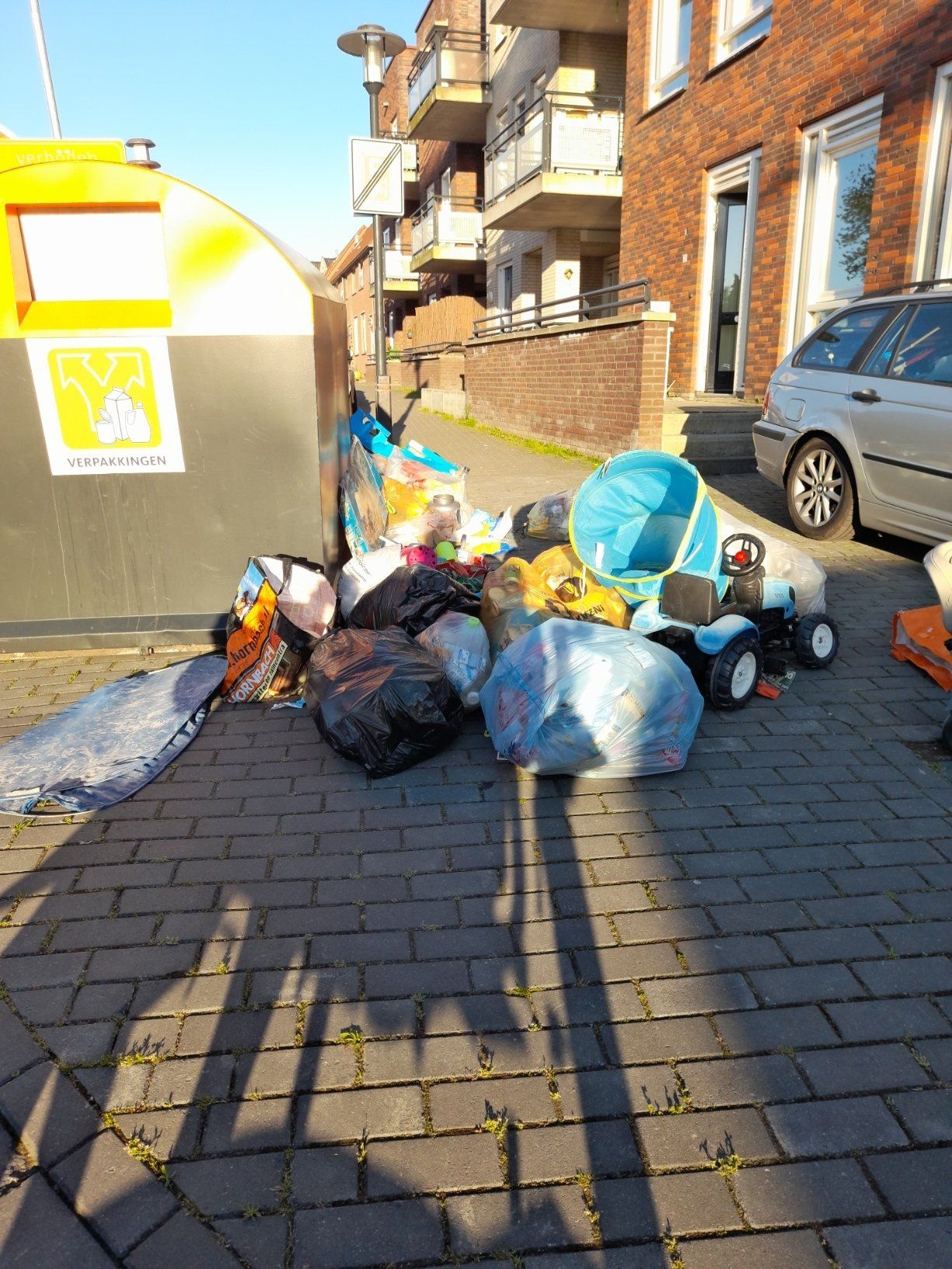 Er ligt een hoop afval op een stoep van bakstenen, naast een afvalcontainer.