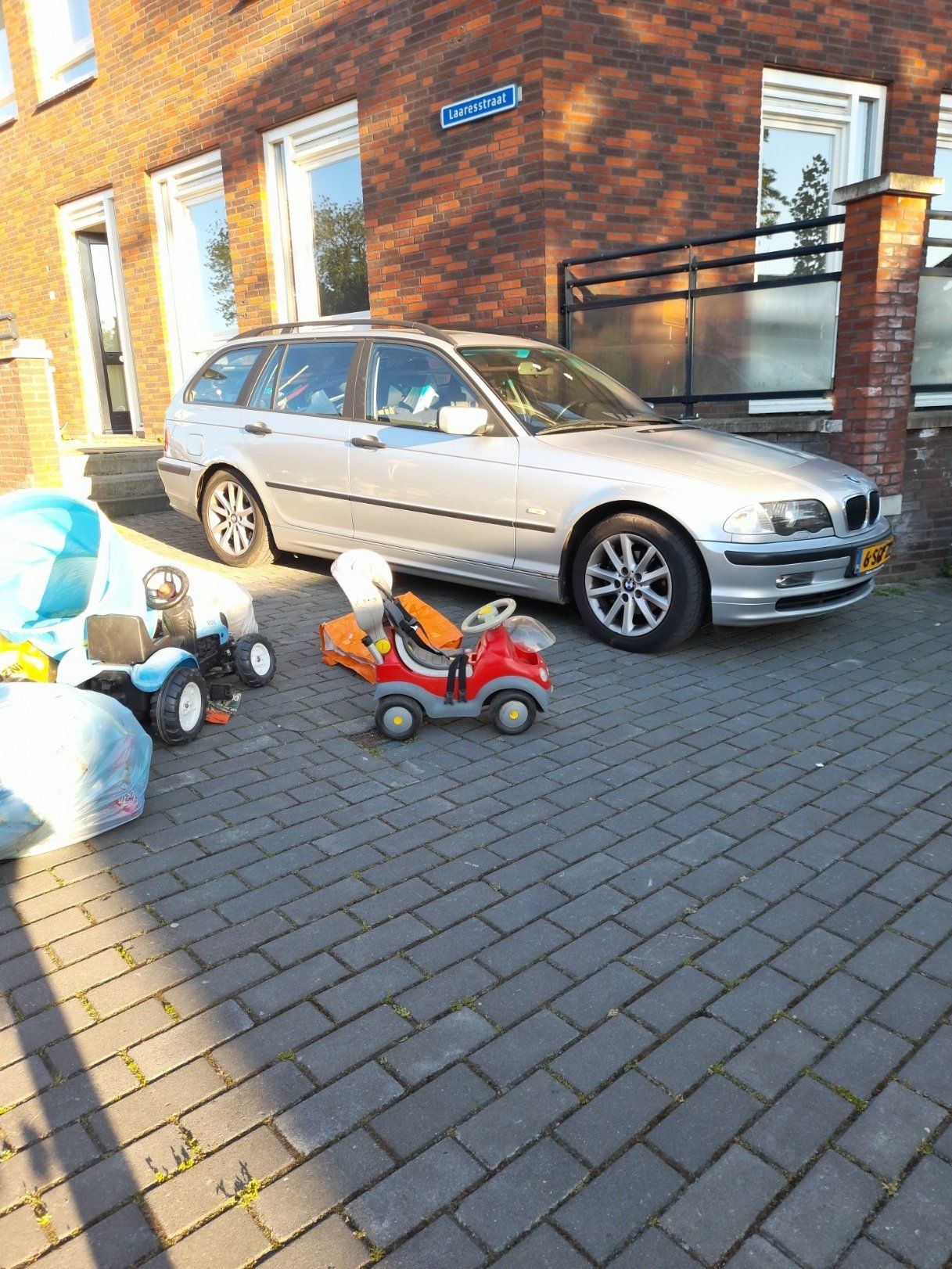 Een zilveren auto staat geparkeerd op een bakstenen oprit naast twee speelgoedauto's.