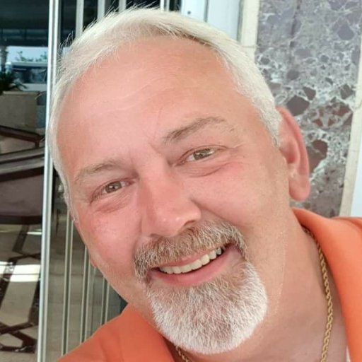 Een man met een baard lacht en draagt een oranje shirt.