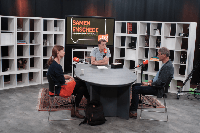 Drie mensen zitten aan een tafel voor een scherm met de tekst samen enschede