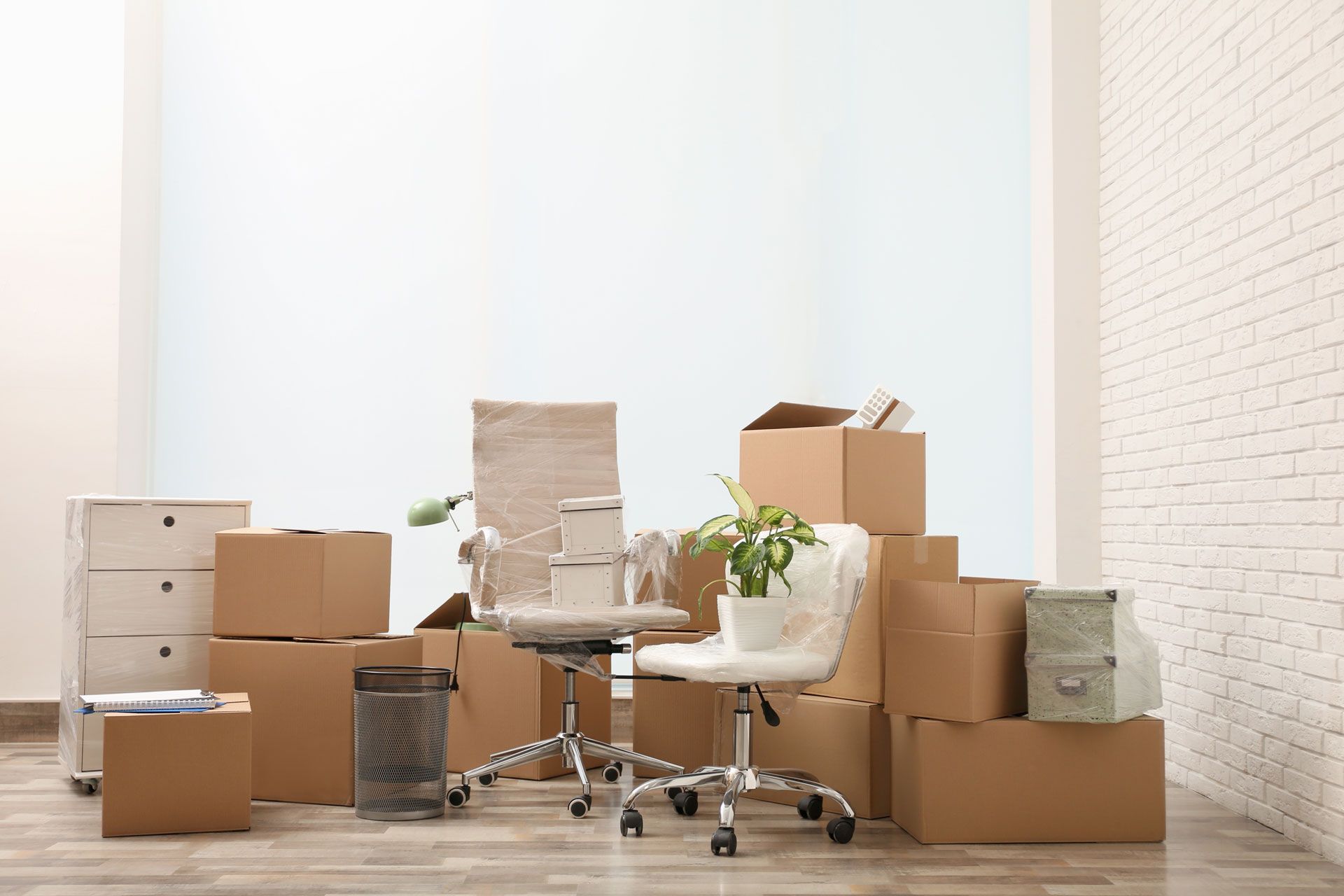 Boxes and office furniture in a bright, empty room; white brick wall; moving.