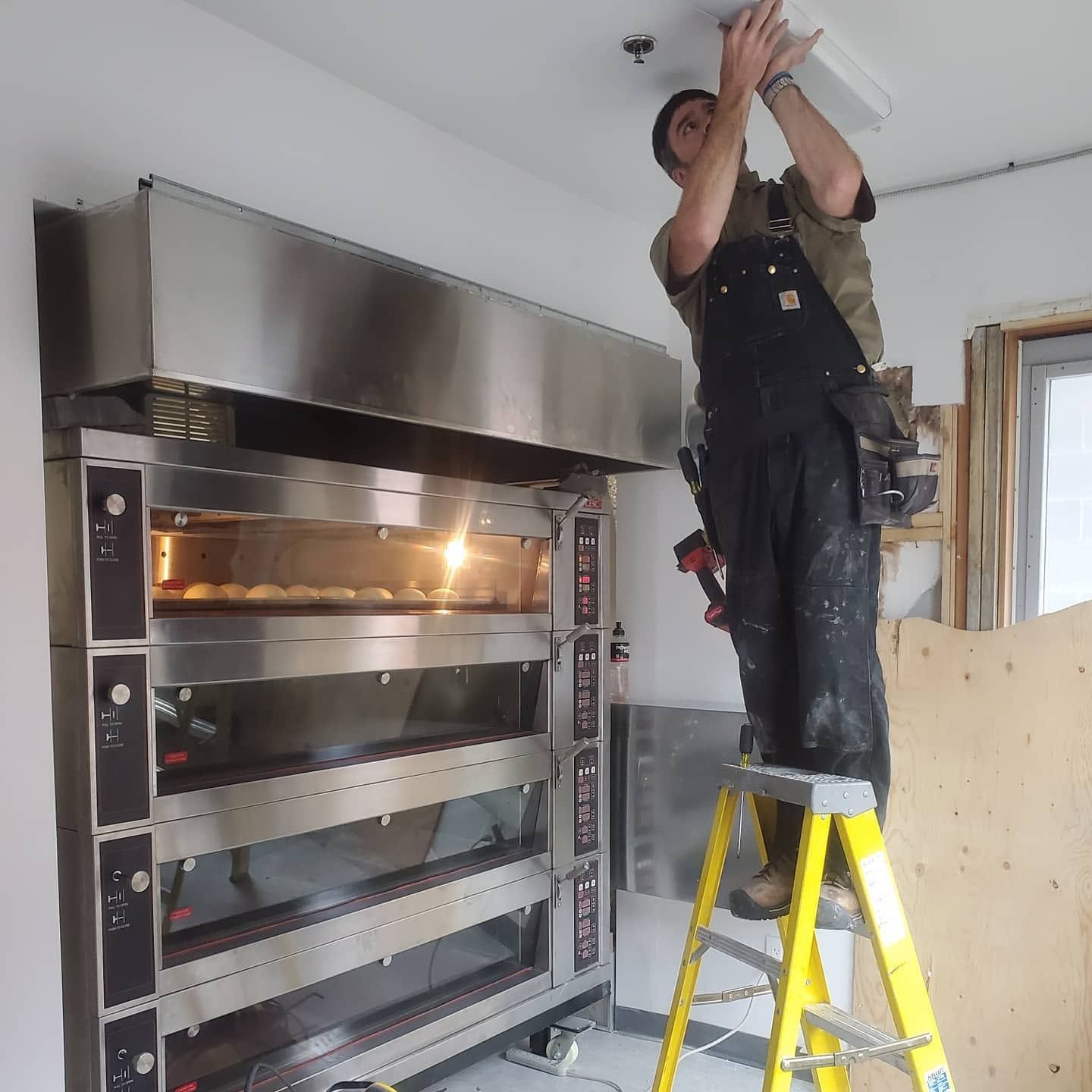 Worker on a yellow ladder installing a ceiling fixture above a large metal oven in a room.