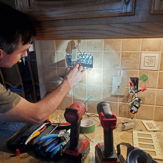 Person wiring an electrical outlet in a tiled kitchen backsplash with tools on the counter.